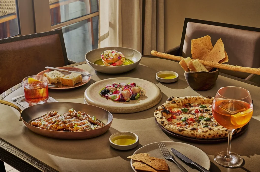 A dining table with various Italian dishes: a pizza, a pasta dish, salad, bread with oil, and a cocktail, set in a cozy, sunlit room.