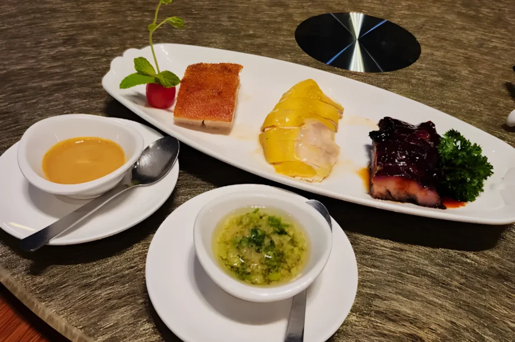 A gourmet plate with roasted meats: crispy pork belly, yellow chicken, and glazed pork, garnished with greens. Two sauces in bowls accompany the dish.