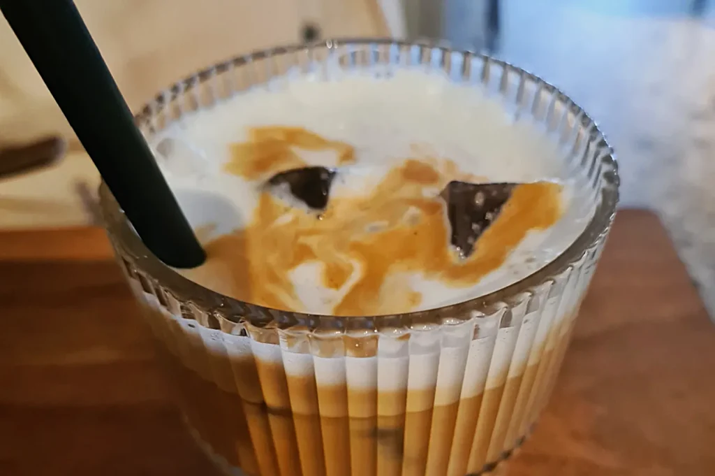 A glass with a ridged texture, filled with iced coffee. There's a layer of light frothy cream on top and visible ice cubes, with a black straw inserted.