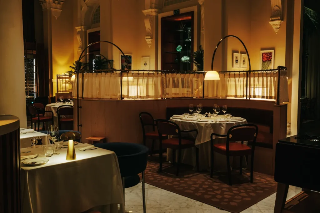Elegant restaurant with dim lighting features round tables set with white tablecloths and wine glasses. Cozy ambiance with modern decor and warm tones.