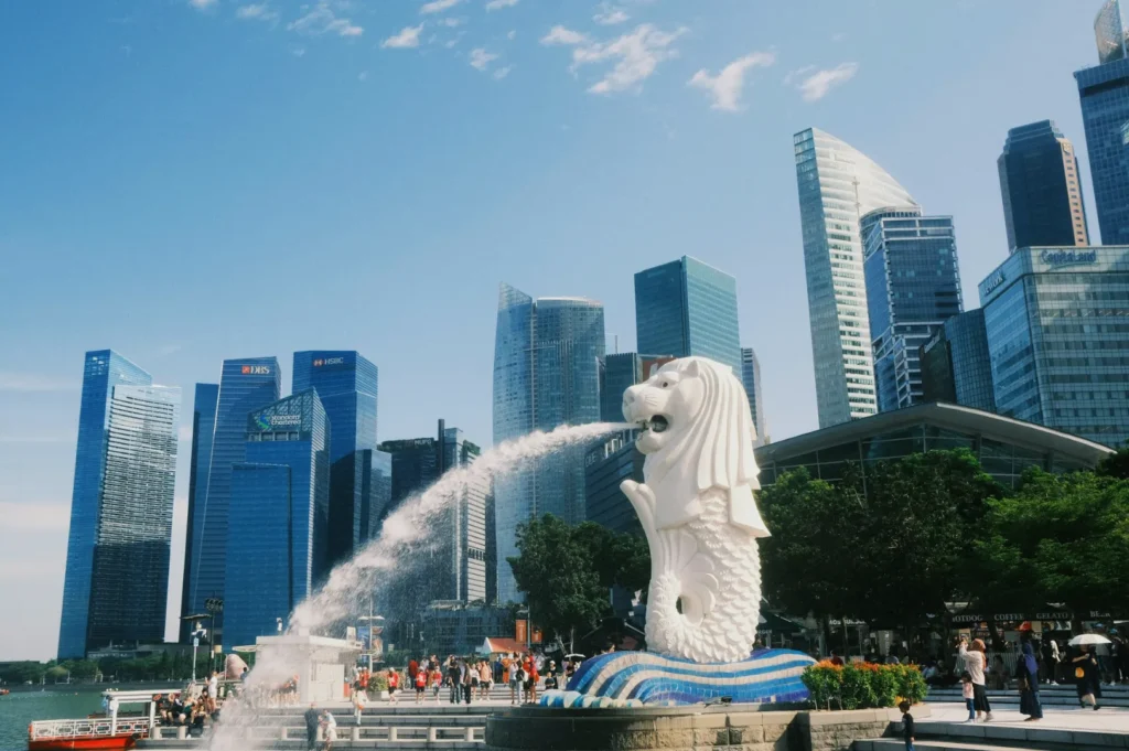 The iconic Merlion statue in Singapore sprays water with a backdrop of modern skyscrapers and a bustling crowd, under a clear blue sky.