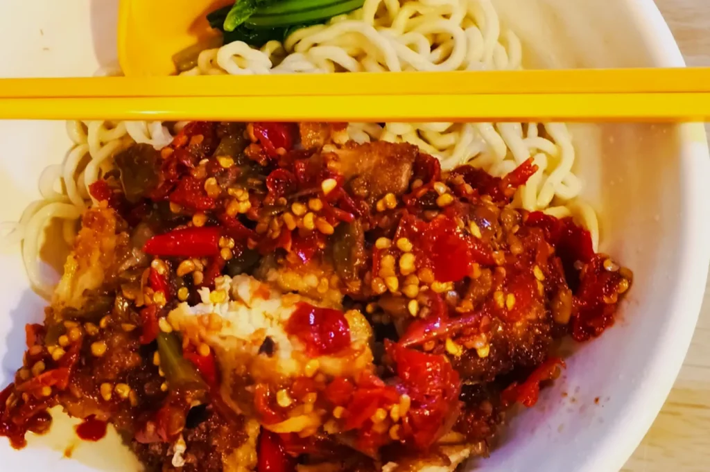 A bowl of noodles topped with spicy red chili sauce and crispy fried chicken, garnished with green vegetables. Bright yellow chopsticks rest on top.