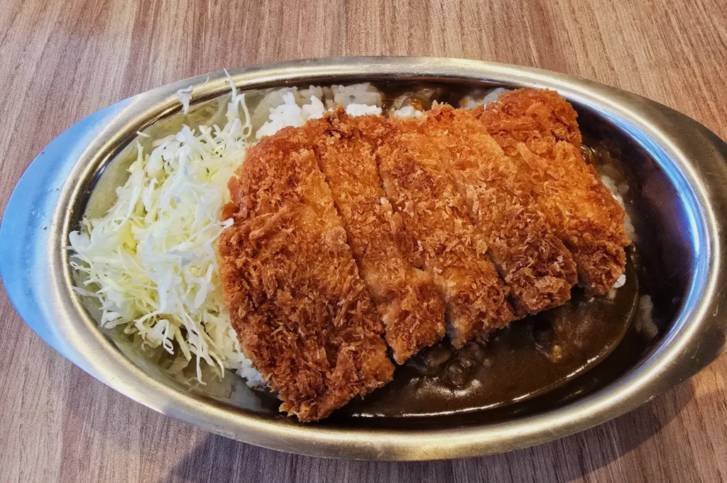 A plate of Japanese katsu curry features crispy breaded pork cutlet, sliced on top of rich curry sauce, white rice, and shredded cabbage.