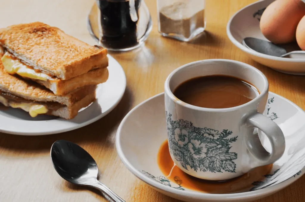 A cup of coffee sits on a saucer, with toast and butter on a plate nearby. A spoon, eggs, and condiment jars are on the wooden table, suggesting a cozy breakfast scene.