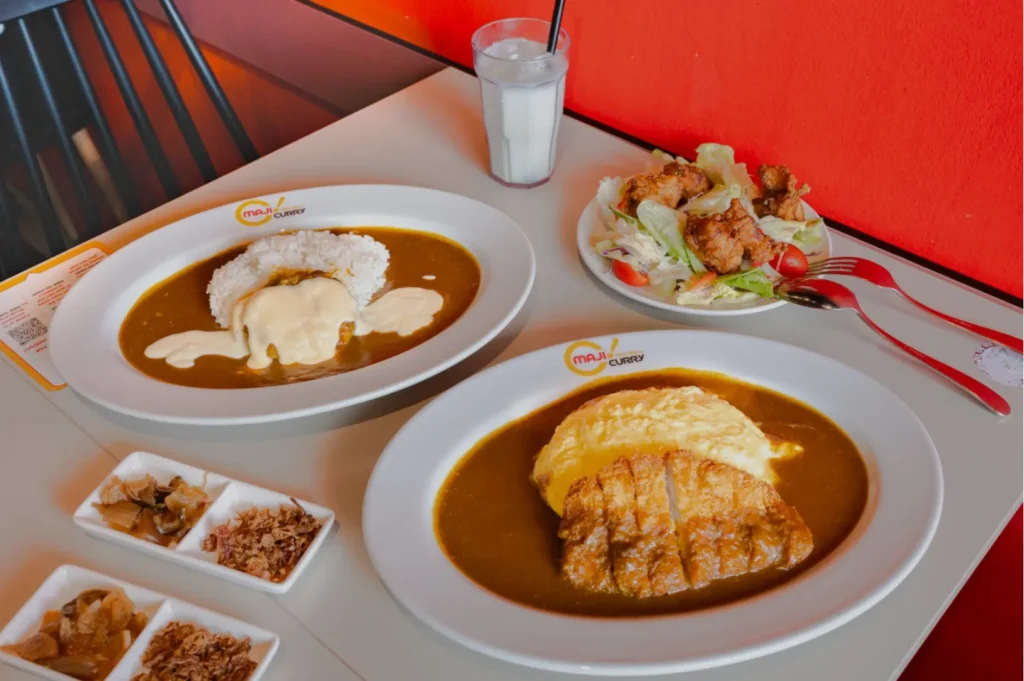 Plates of Japanese curry with rice and omelet, crispy chicken, and salad. Condiments are on the side, and a glass of milk is nearby. Cozy, inviting setting.