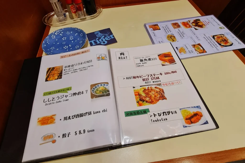 A Japanese restaurant menu on a table shows various dishes with images, prices, and descriptions. Nearby are condiments and a blue plate, creating a cozy, inviting dining setup.