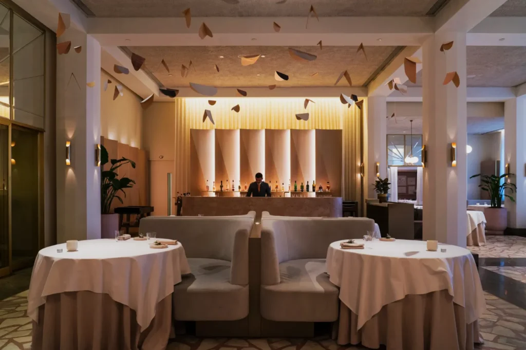 Cozy restaurant interior with round tables and beige booths, each covered with white tablecloths. A bartender stands behind a softly lit bar in the background. Abstract mobiles hang from the ceiling, enhancing the elegant atmosphere.