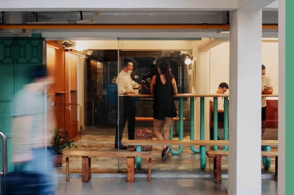 A modern cafe scene with glass walls showing two people standing inside, engaged in conversation. Others are seated, absorbed in their phones. The decor is minimalistic and bright, creating a lively, urban atmosphere.
