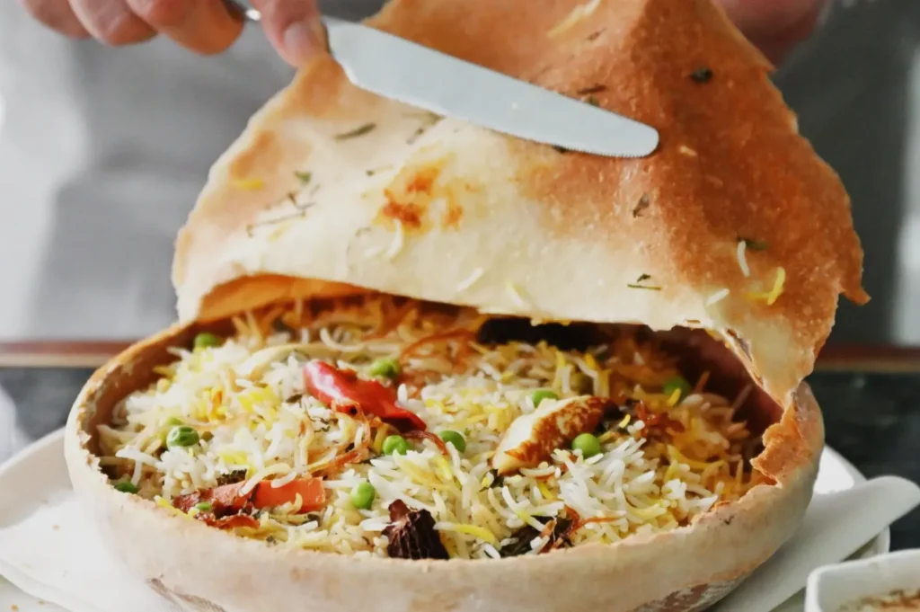 A hand lifts a crispy naan crust off a bowl of colorful biryani rice, revealing vibrant vegetables and spices within, creating a warm, inviting atmosphere.