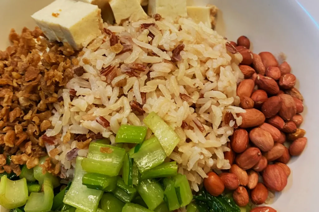 A bowl of white and red rice surrounded by chopped green vegetables, cubed tofu, roasted peanuts, and minced garlic, creating a fresh, wholesome feel.