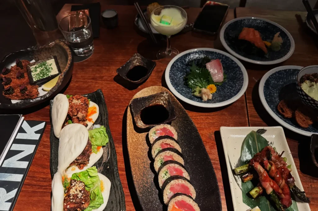 A wooden table is filled with various Asian dishes, including bao buns, sushi rolls, sashimi, grilled skewers, and dipping sauces, creating a vibrant dining experience.