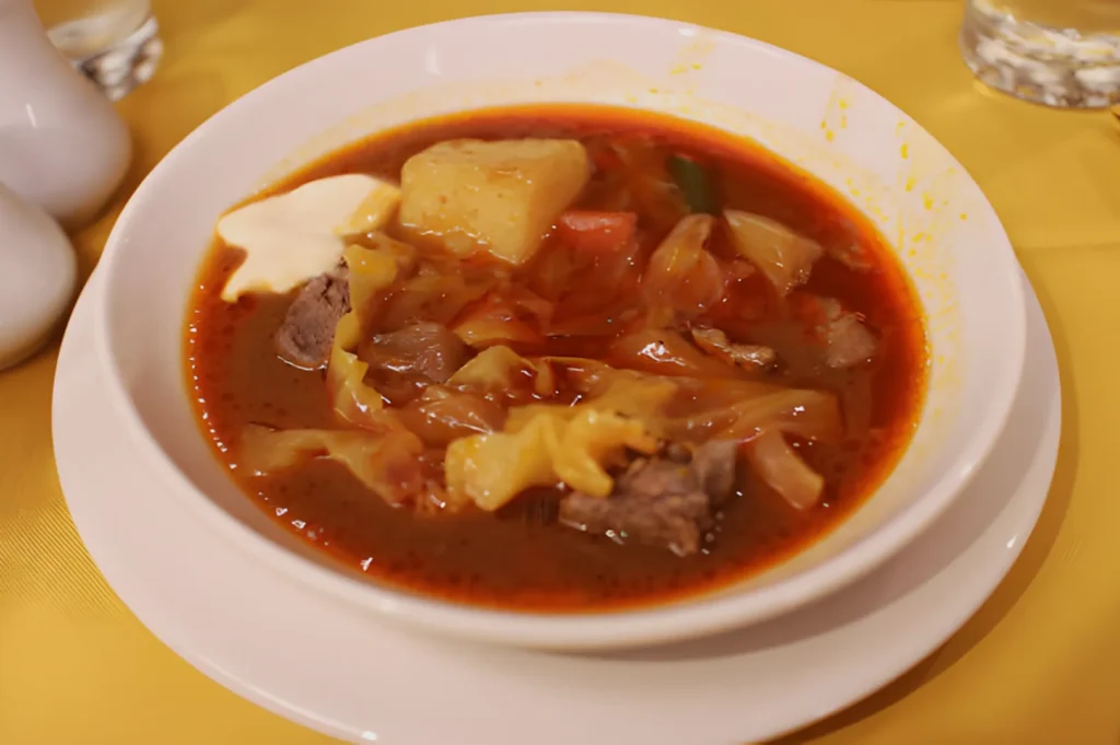 A bowl of hearty red soup with chunks of beef, cabbage, potato, and a dollop of sour cream, set on a yellow tablecloth, creating a warm and inviting mood.