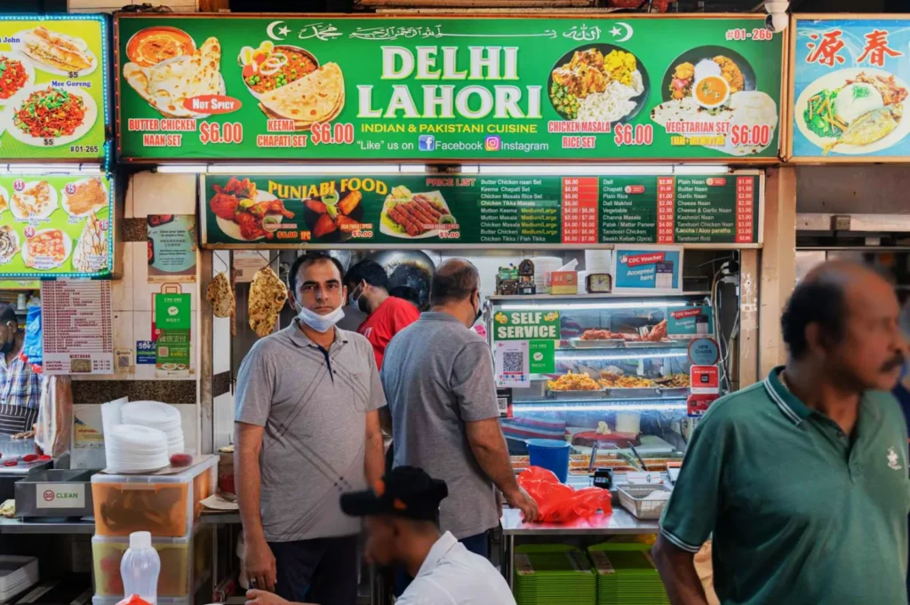 Bustling food stall labeled "Delhi Lahori" offers Indian & Pakistani cuisine. Customers gather around, menu displays vibrant dishes and prices, lively market vibe.
