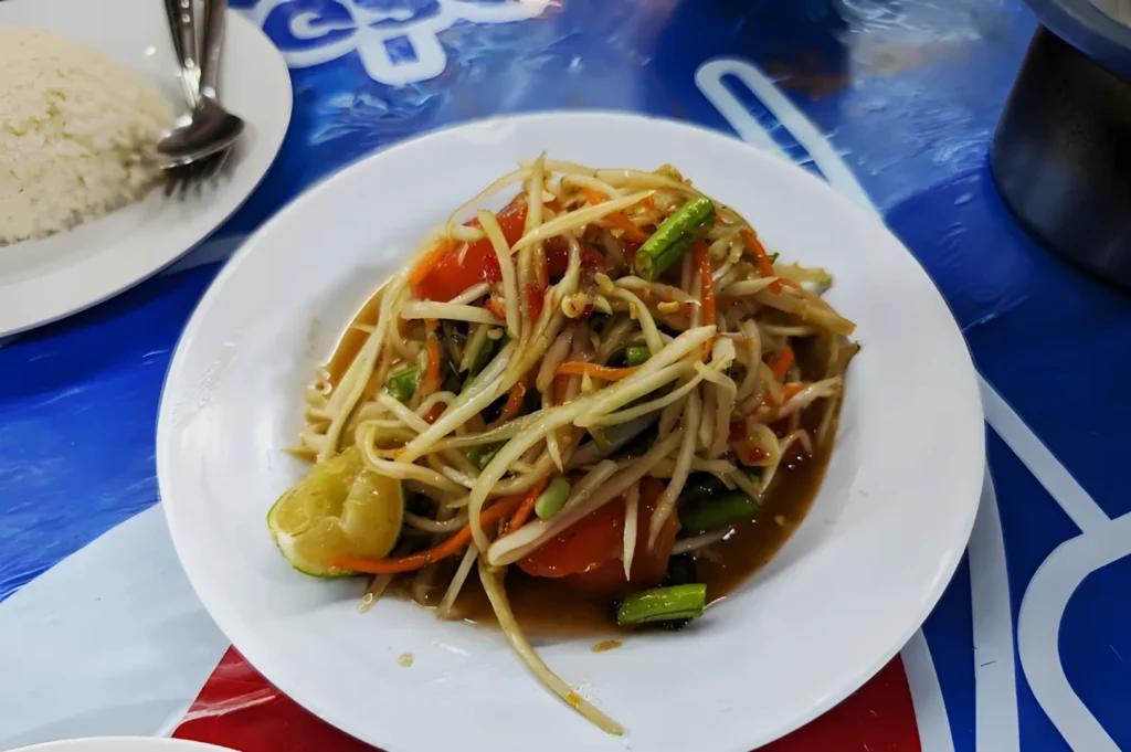 A vibrant Thai papaya salad on a white plate, featuring shredded green papaya, tomatoes, lime, and green beans. A side plate holds rice and utensils.