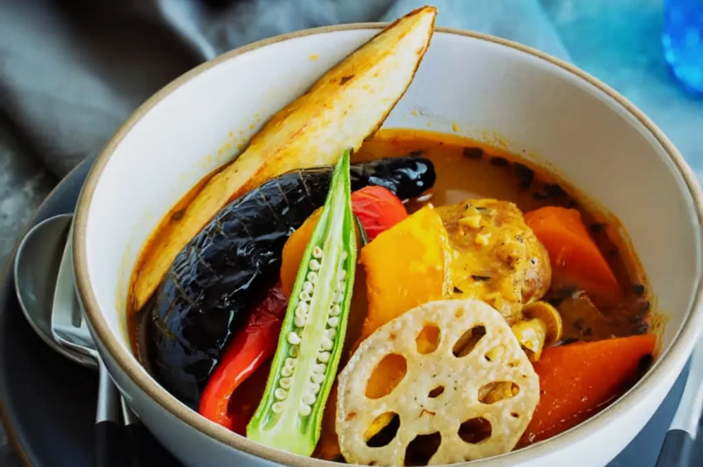 A bowl of vibrant vegetable curry features eggplant, okra, bell peppers, lotus root, and carrot in a rich, spiced broth. The mood is warm and hearty.