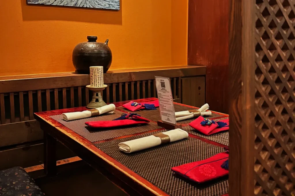 Cozy dining setup with a rustic wooden table, red napkins, and white cloth napkins neatly arranged. Warm lighting and an artistic ceramic pot add charm.