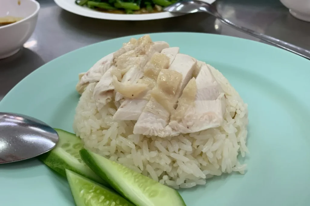 Plate of Hainanese chicken rice with sliced chicken on steamed rice, garnished with cucumber slices. A spoon is beside; a dish in the background.