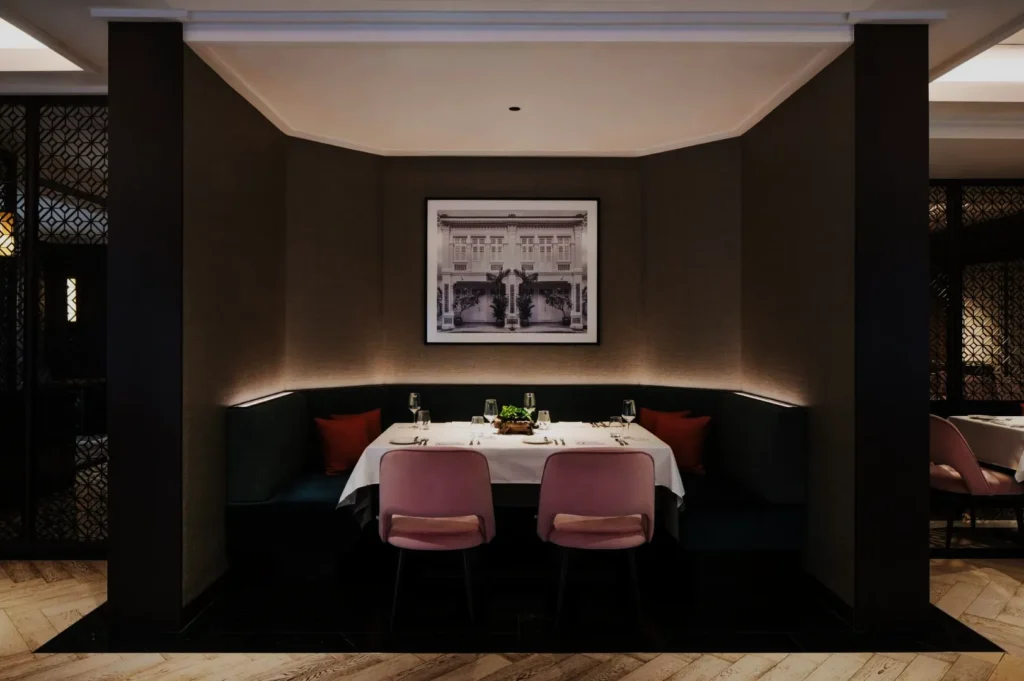 Elegant restaurant booth with pink chairs and teal seating, set with glasses and cutlery. A framed black-and-white photo decorates the neutral walls.