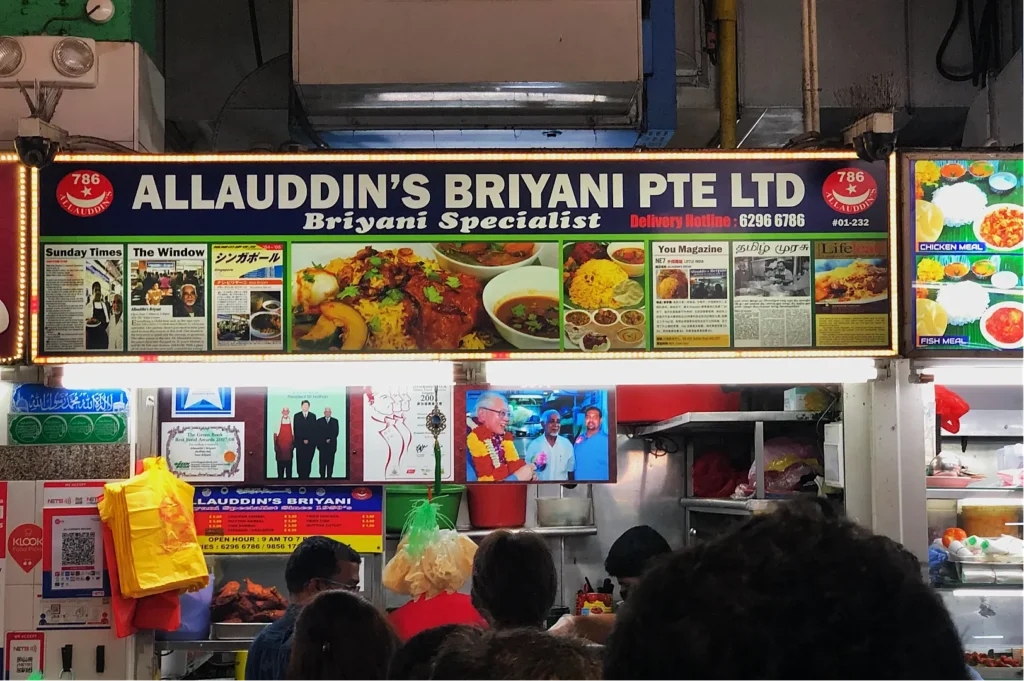 Signage for Allauddin’s Briyani Pte Ltd at a food stall. The sign features images of briyani dishes and newspaper clippings, indicating accolades. Below are people waiting to order, creating a bustling and lively atmosphere.