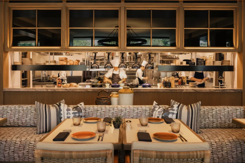 Cozy restaurant with an open kitchen design; chefs in white hats cook lively dishes. The foreground features a set dining table with striped cushions.