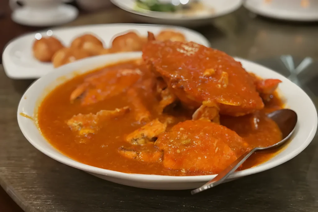Plate of chili crab in rich, red sauce with a spoon on the side. In the background, a dish with round fried buns. Warm and savory setting.