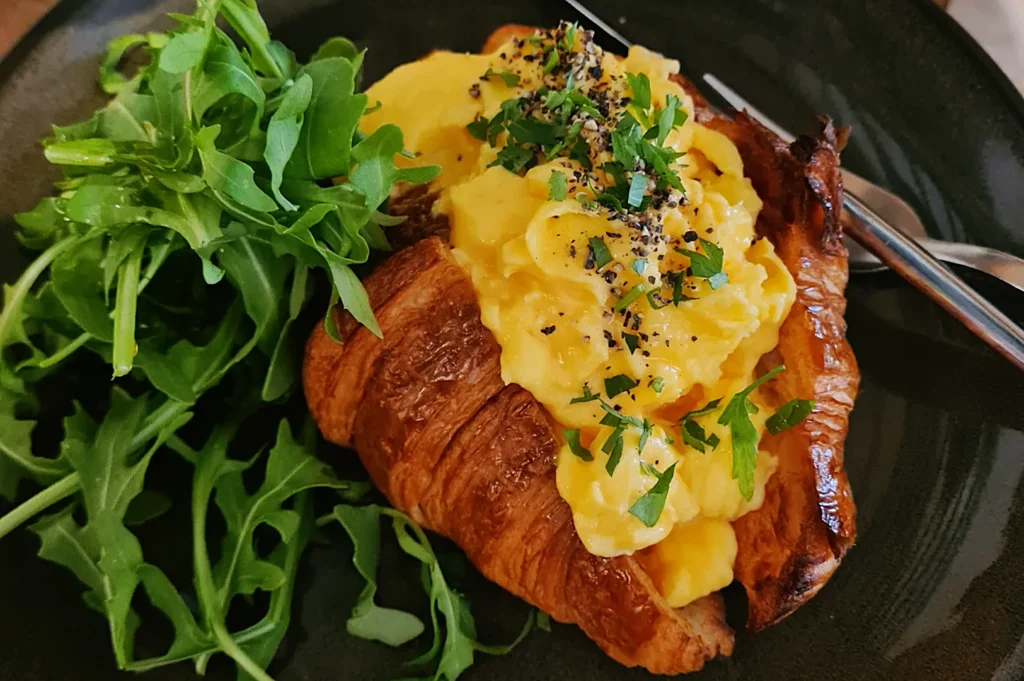 A croissant topped with creamy scrambled eggs, garnished with fresh herbs and black pepper, accompanied by a small side of fresh arugula.