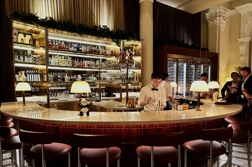 A stylish bar with warm lighting and a semi-circular marble counter. Bartender serves drinks; patrons chat. Bottles line the shelves, creating a cozy vibe.