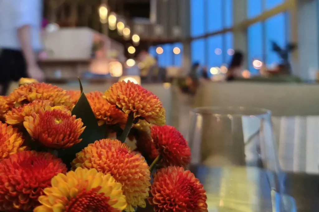 A bouquet of vibrant orange and red flowers in focus on a restaurant table, with a softly blurred background showing warm lights and large windows.