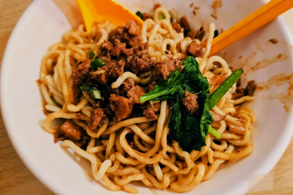 A bowl of noodles topped with chunks of meat and green leafy vegetables. A yellow spoon sticks out from the bowl, hinting at a casual dining scene.