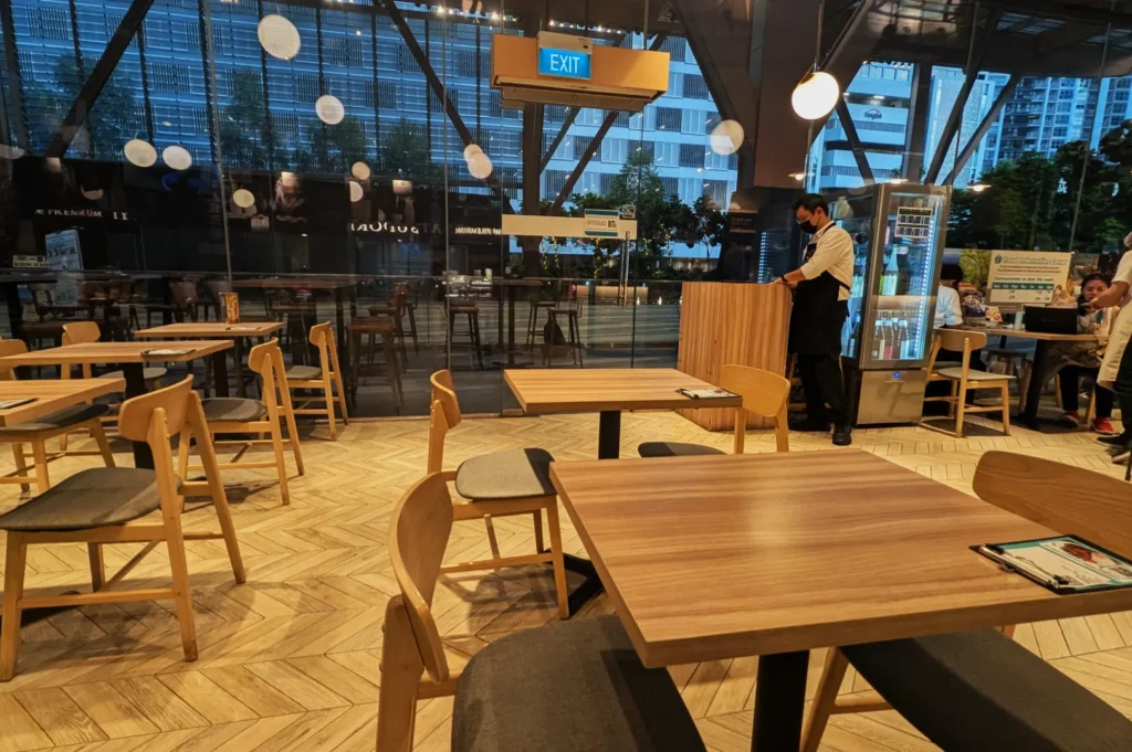 Modern café interior with wooden tables and chairs, large windows, and a herringbone floor. A staff member stands near the counter, creating a welcoming and calm atmosphere.