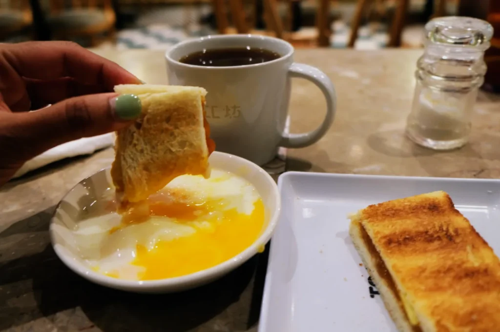 A hand dips toast into a runny half-boiled egg in a bowl. Nearby, there's a mug of tea and a plate with more toast, conveying a cozy breakfast scene.