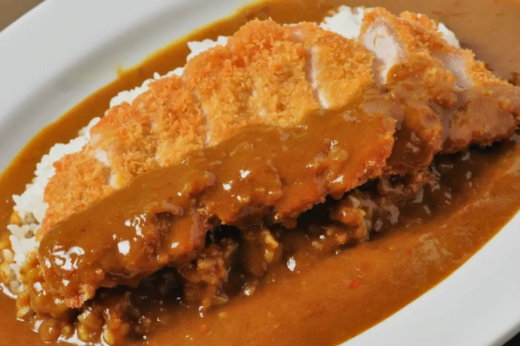 A plate of Japanese curry rice topped with crispy breaded katsu, drizzled with rich brown curry sauce. The dish appears warm and savory.