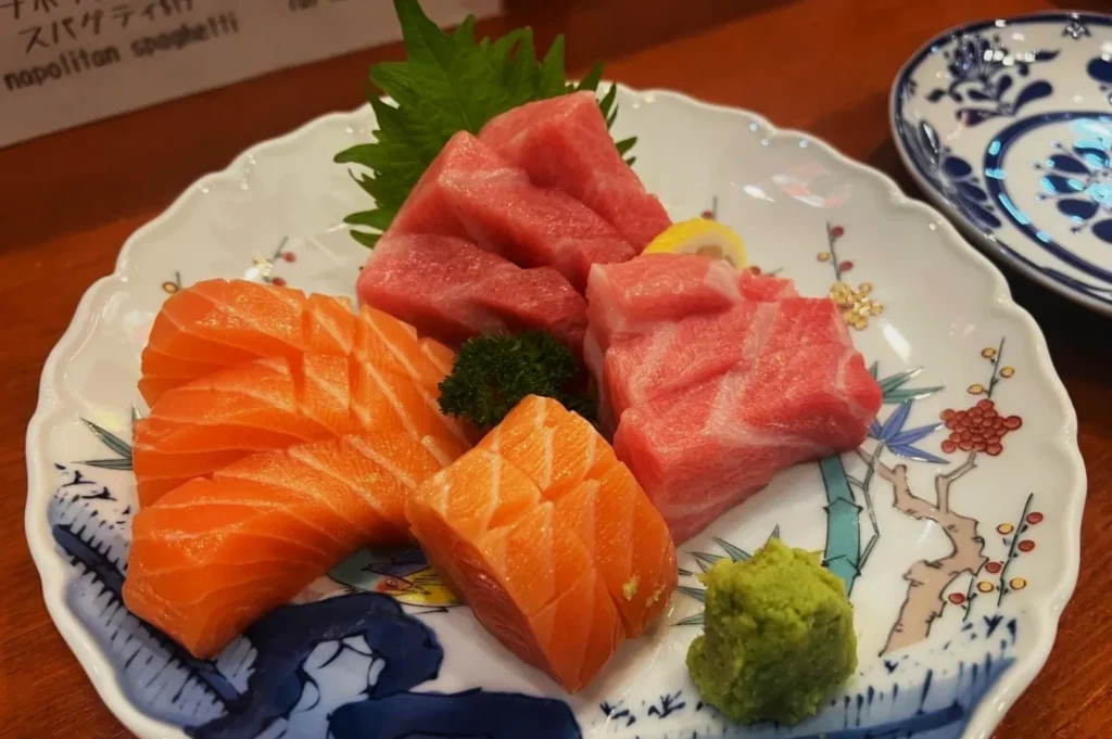 Assorted sashimi on a decorative plate includes vibrant salmon and tuna slices, garnished with green leaves, parsley, and a wasabi dollop.