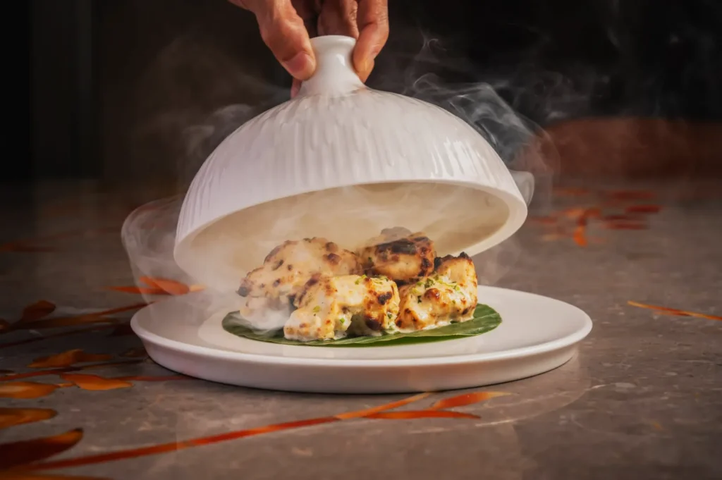 Hand lifting a white cloche to reveal steaming, grilled chicken pieces on a leaf. The dish is on a marble table with a sophisticated ambiance.
