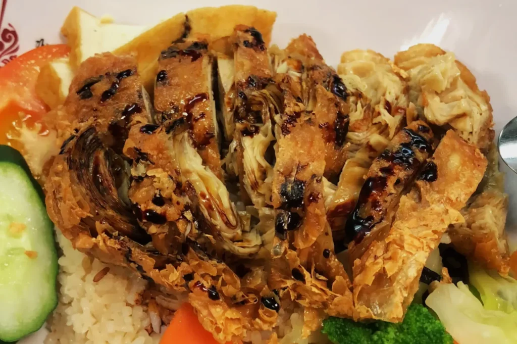 Close-up of a dish featuring sliced, grilled mock meat with a crispy texture, drizzled with dark sauce. Served on rice with cucumber, tomato, and steamed vegetables.