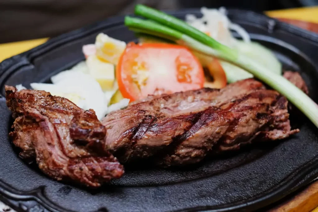 Grilled steak on a dark plate with sliced tomato, onions, and green onions. The dish appears appetizing and freshly prepared.