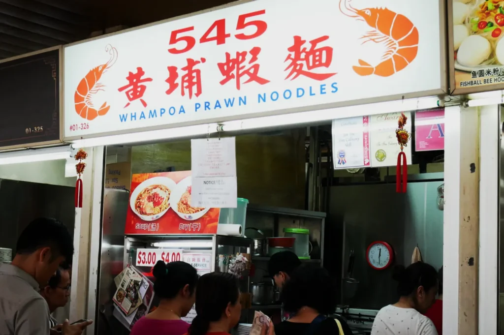 A small, bustling food stall named "545 Whampoa Prawn Noodles" with a sign featuring red prawns. People queue eagerly awaiting their orders.