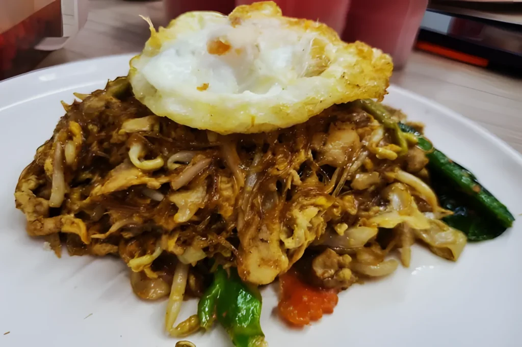A white plate holds stir-fried noodles mixed with vegetables and topped with a sunny-side-up egg, creating a savory, appetizing presentation.