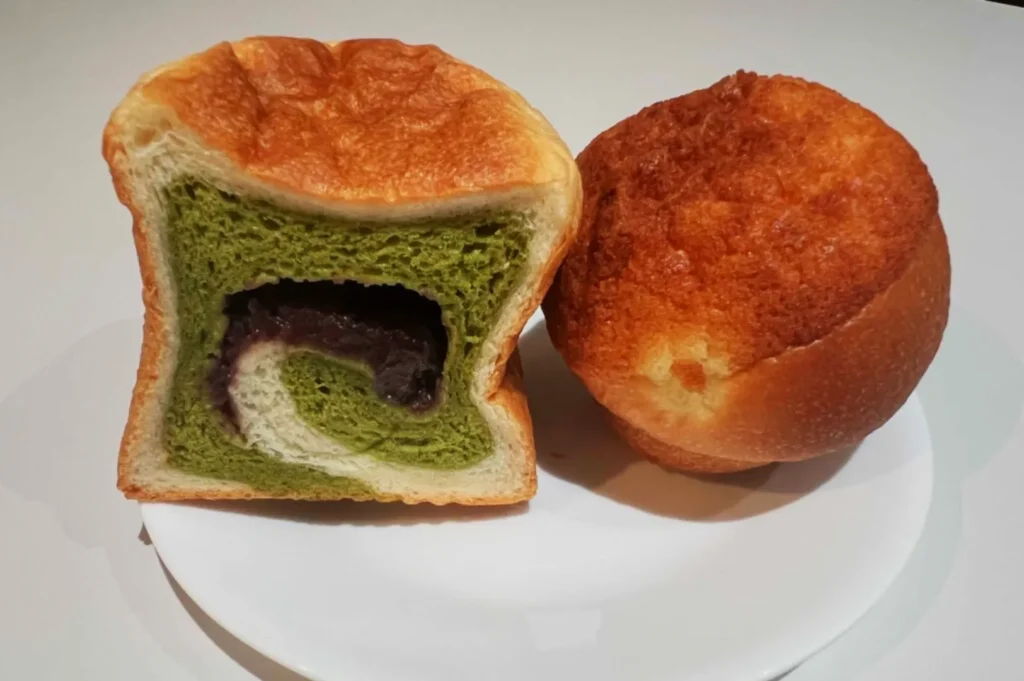 Two breads on a white plate; one sliced revealing green matcha and red bean swirl filling, the other round with a golden crust.