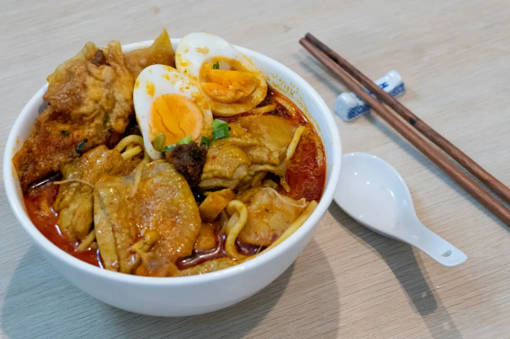 A bowl of spicy noodle soup with sliced hard-boiled egg, fried tofu, and meat chunks. Chopsticks and a soup spoon rest on the side, creating a warm, appetizing setting.