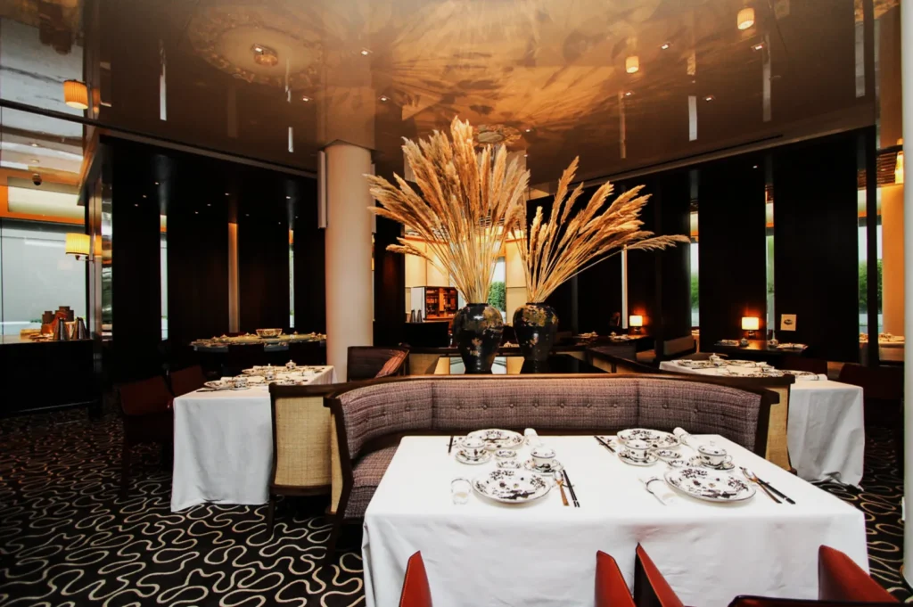 Elegant restaurant interior with round tables set with white linens and intricate china. Tall decorative vases with pampas grass create a sophisticated ambiance.