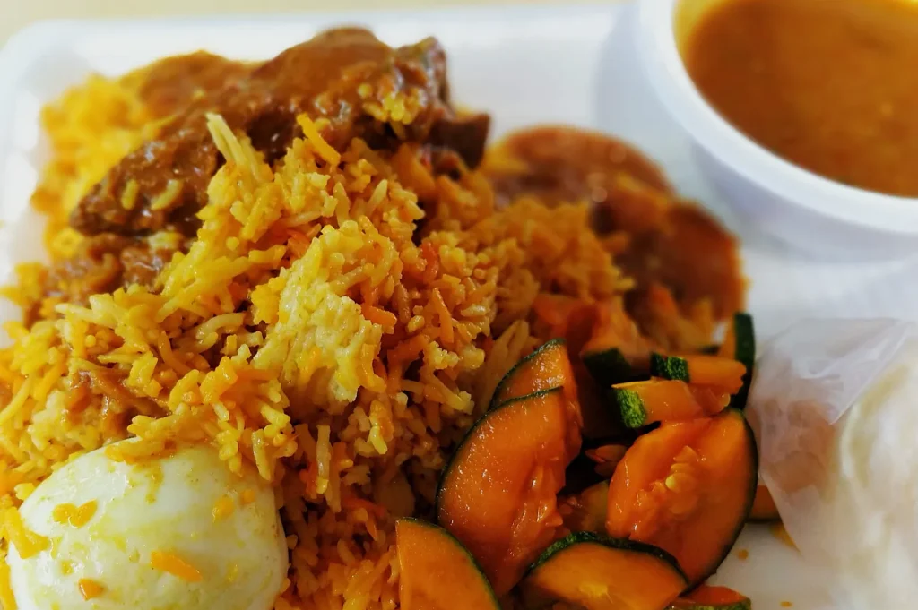 A plate of yellow rice with chicken curry, slices of cooked zucchini, a boiled egg, and a side of broth in a small bowl, creating a warm, hearty meal.