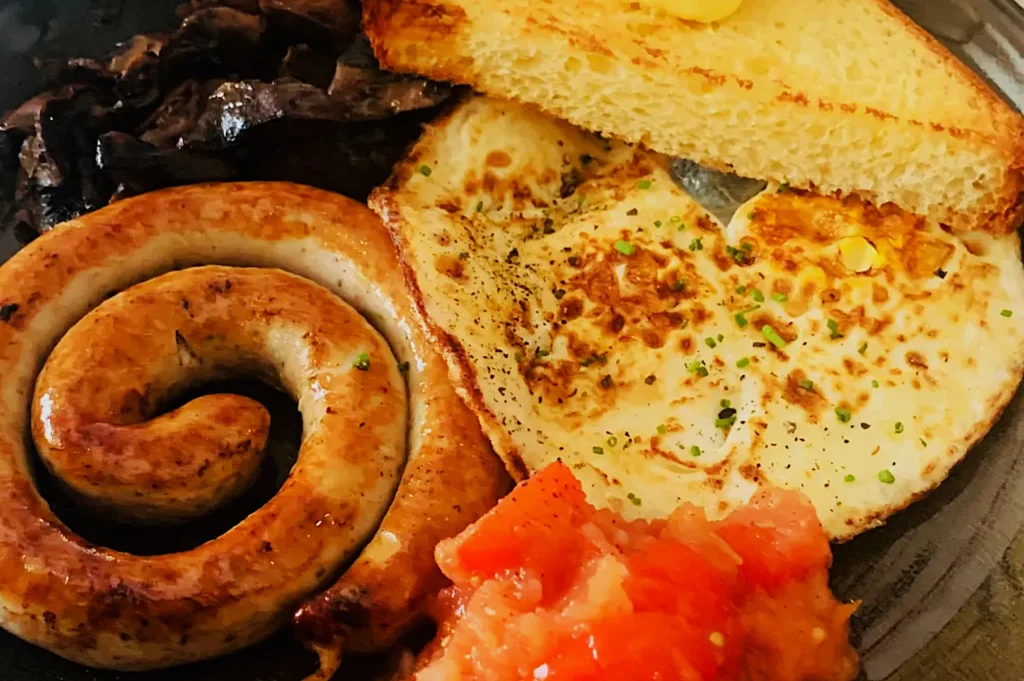 A hearty breakfast plate with a coiled sausage, sunny-side-up egg sprinkled with chives, buttered toast, sautéed mushrooms, and diced tomatoes.