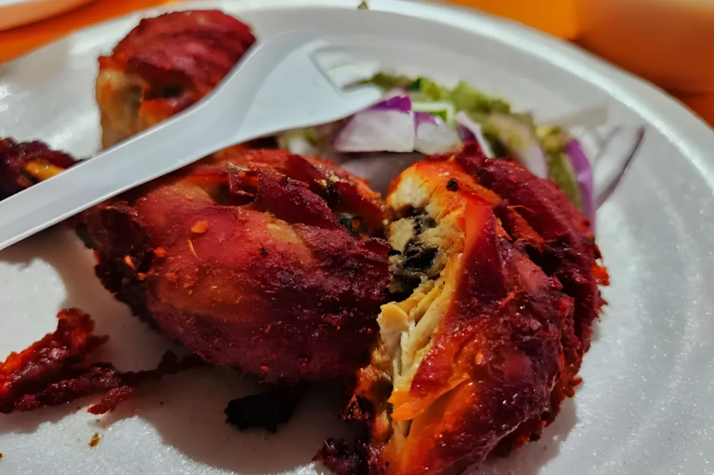 Plate of marinated, cooked meat with a crispy red exterior, garnished with onion slices and salad. A plastic fork rests on the side, creating a casual dining feel.