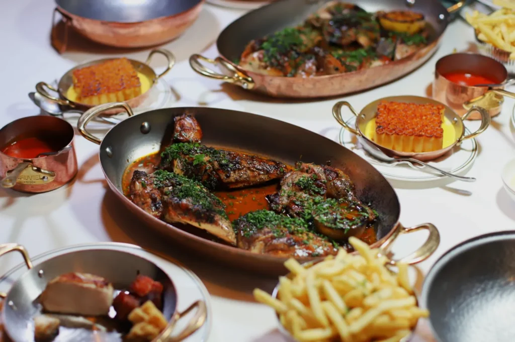 A table set with gourmet dishes, including roasted chicken with herbs, fries, and custard topped with red garnish, all in elegant copper cookware.