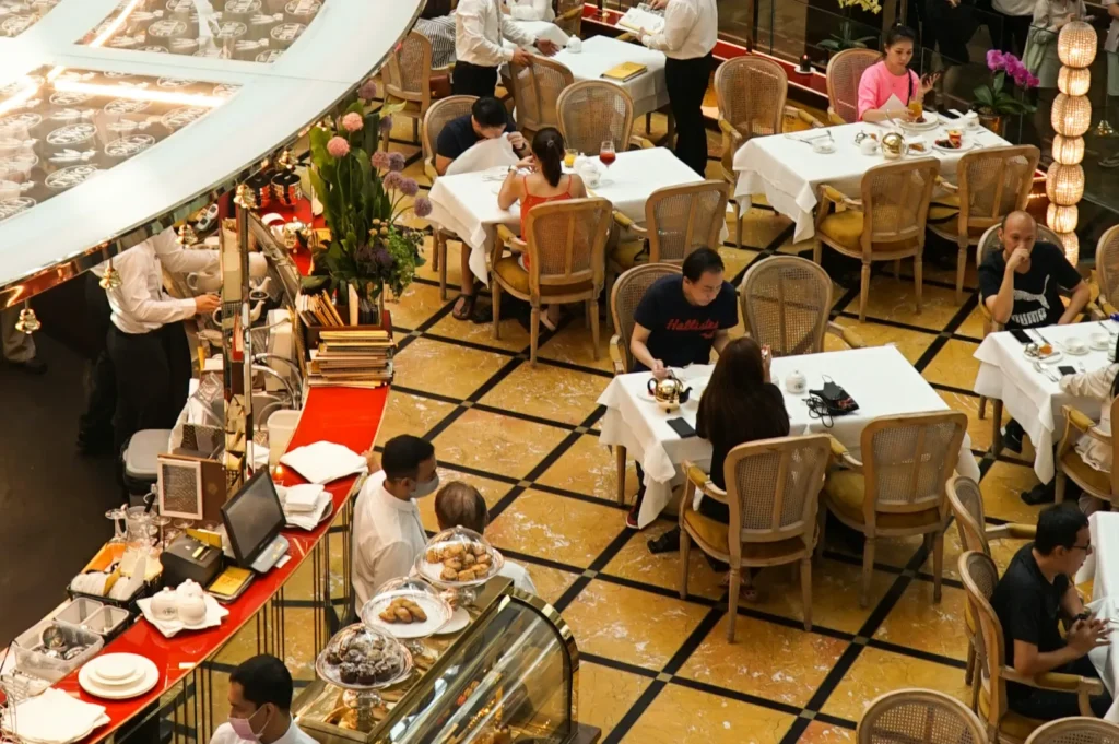 Elegant cafe scene with people dining at tables covered in white cloths. Waitstaff serve guests amid warm lighting and floral decorations. Airy, bustling ambiance.