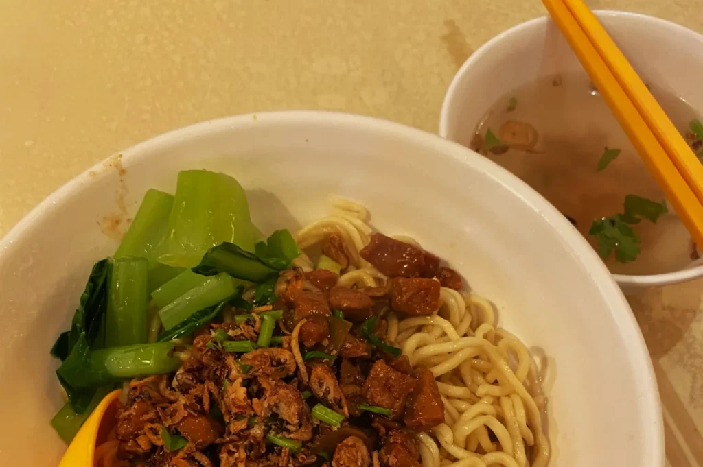 Bowl of noodles with diced meat and green vegetables, accompanied by a side of clear broth. Yellow chopsticks rest on the broth bowl, creating a cozy dining vibe.