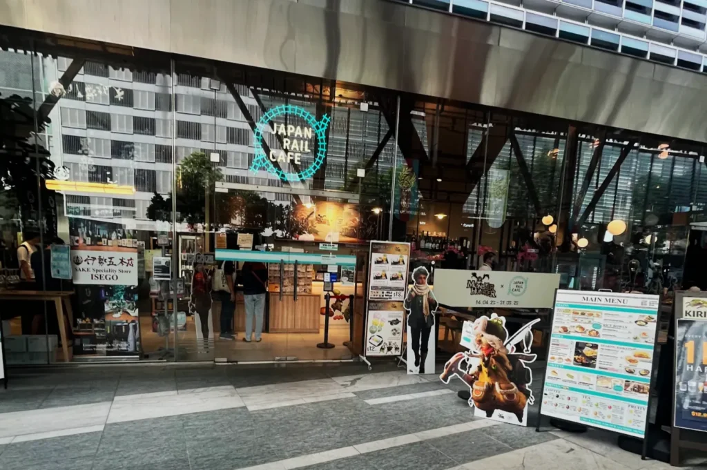 Front view of Japan Rail Cafe with large glass doors, colorful menu boards, and anime character cutouts. The cafe interior is warmly lit.