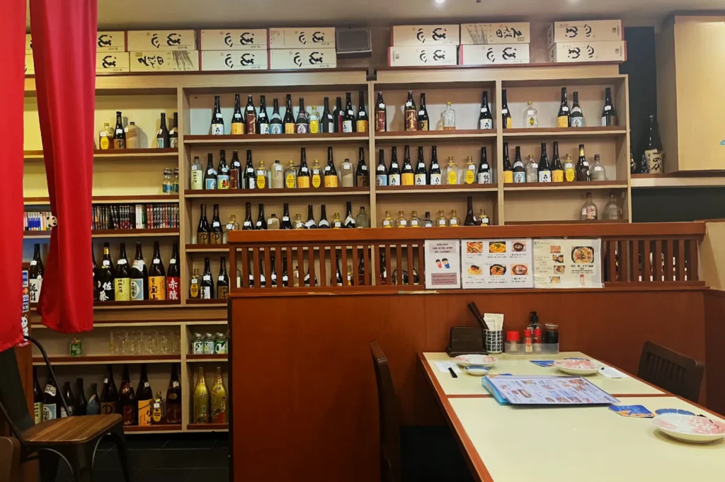 Cozy Japanese restaurant interior with shelves of sake bottles and boxes, a table with menus and utensils, and warm wooden tones, conveying a welcoming ambiance.