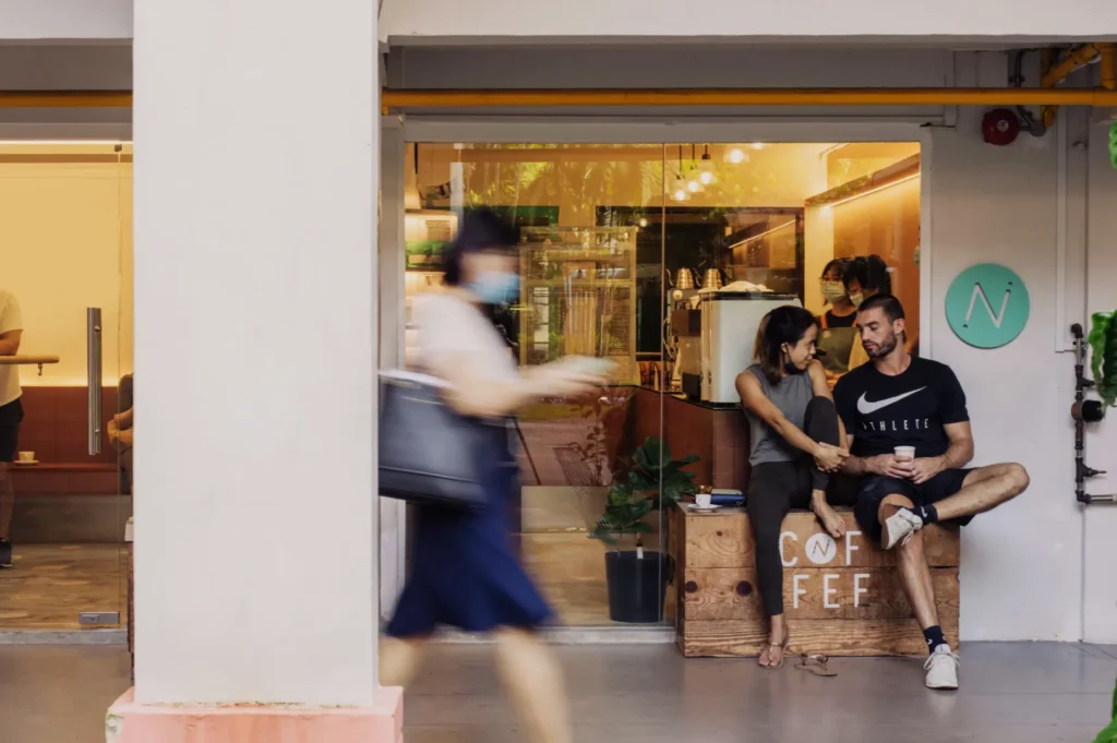 A couple sits on a bench outside a cozy café, chatting with a relaxed demeanor. A blurred pedestrian walks by, conveying movement and urban life.